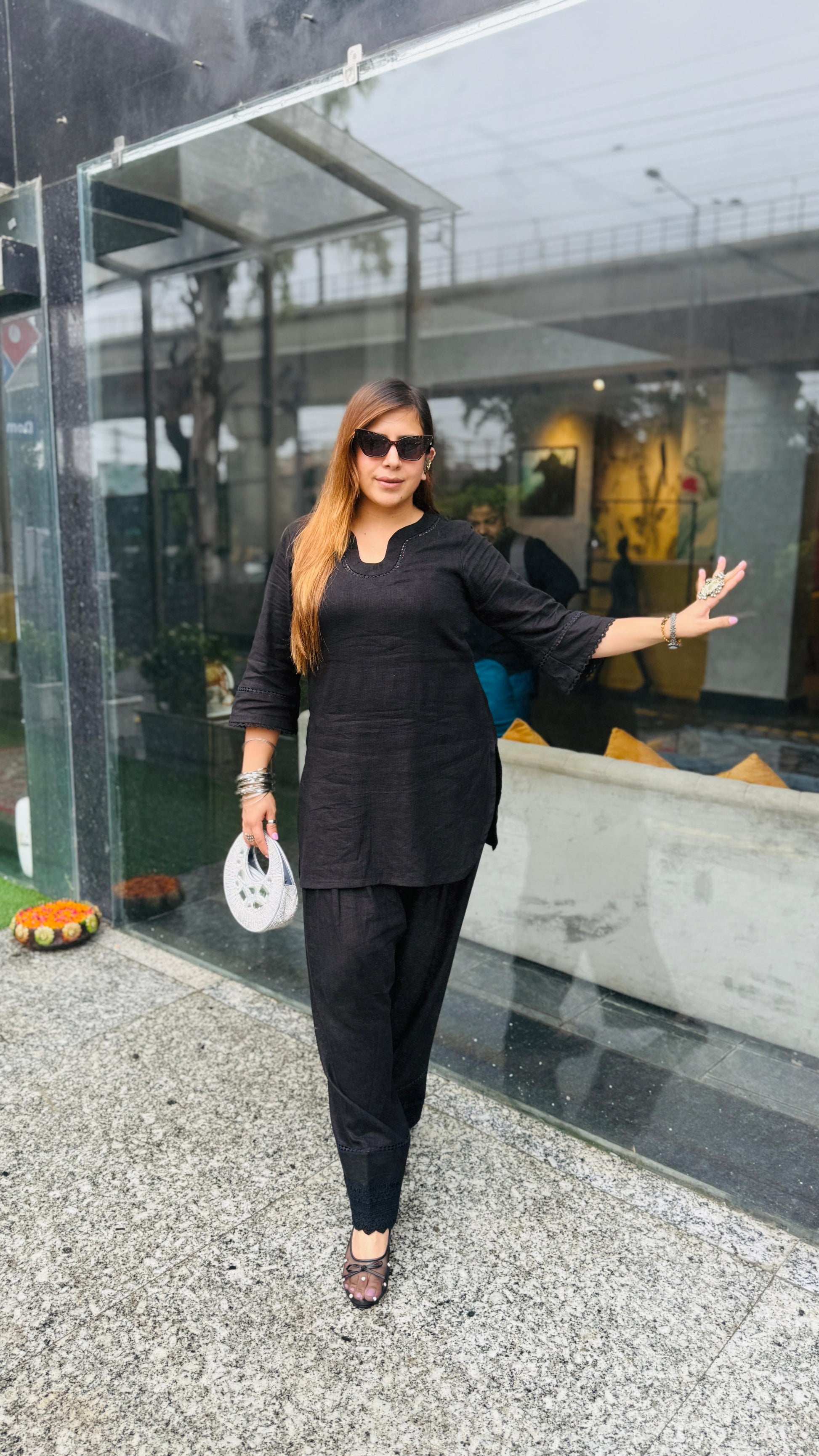 A woman wearing a black three-piece kurta set with sunglasses and holding a small white bag, posing in front of a modern glass storefront.