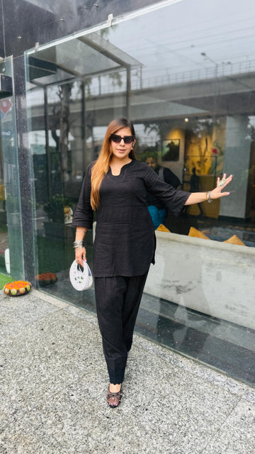 A woman wearing a black three-piece kurta set with sunglasses and holding a small white bag, posing in front of a modern glass storefront.
