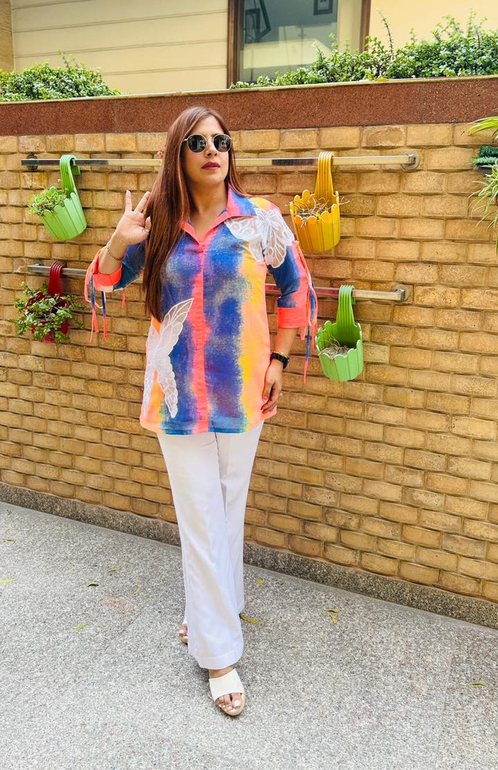 Woman in a colorful shirt and white pants standing in front of a wooden wall with plants.