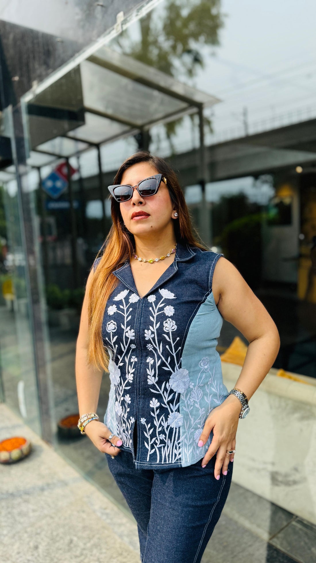 A woman walking outdoors in front of a modern glass building, wearing a sleeveless top with a blue and white patterned yoke and dark blue flared pants.