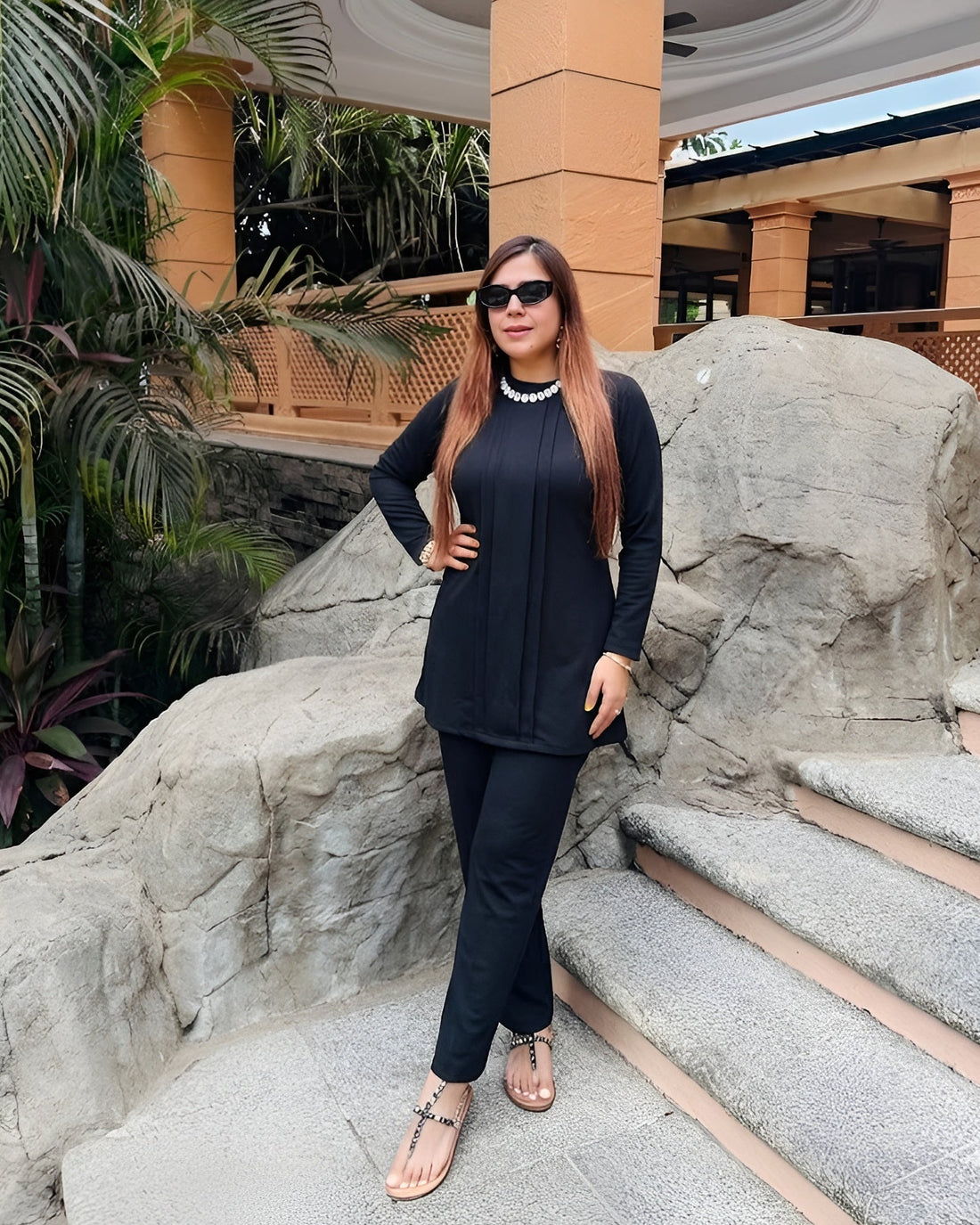 Woman in black outfit standing on stone steps with greenery and architectural elements in the background