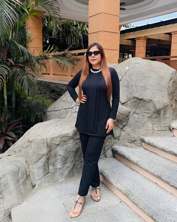 Woman in black outfit standing on stone steps with greenery and architectural elements in the background