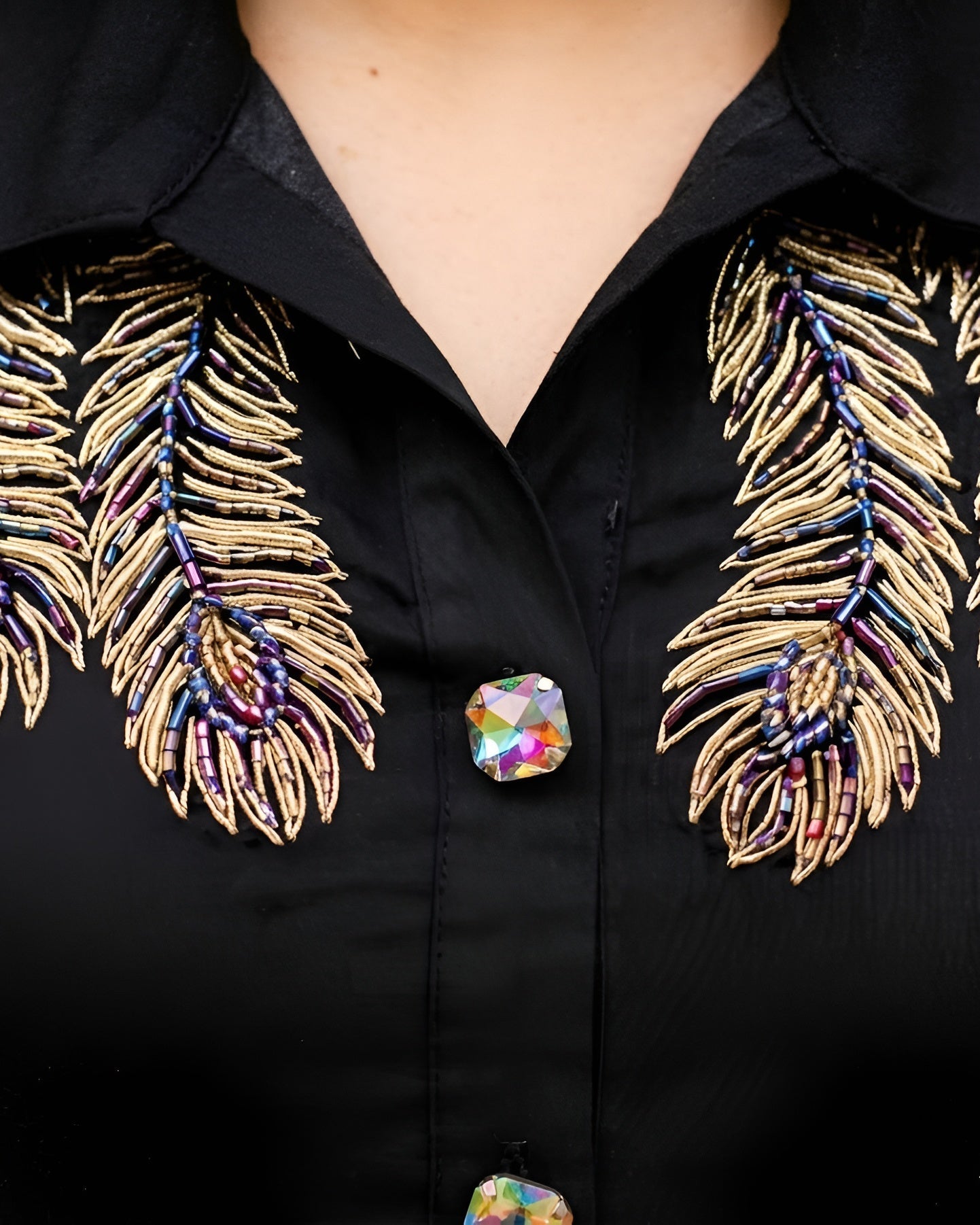 Close-up of a black button-front collared top with large embroidered feather motifs in metallic gold, blue, and purple on both sides of the chest, plus iridescent decorative buttons.