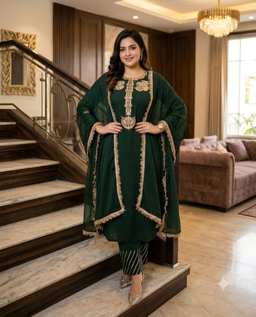 A woman models a dark green traditional Indian outfit with gold embroidery, standing on marble stairs in a luxurious home interior with a staircase, sofa, and chandelier visible.