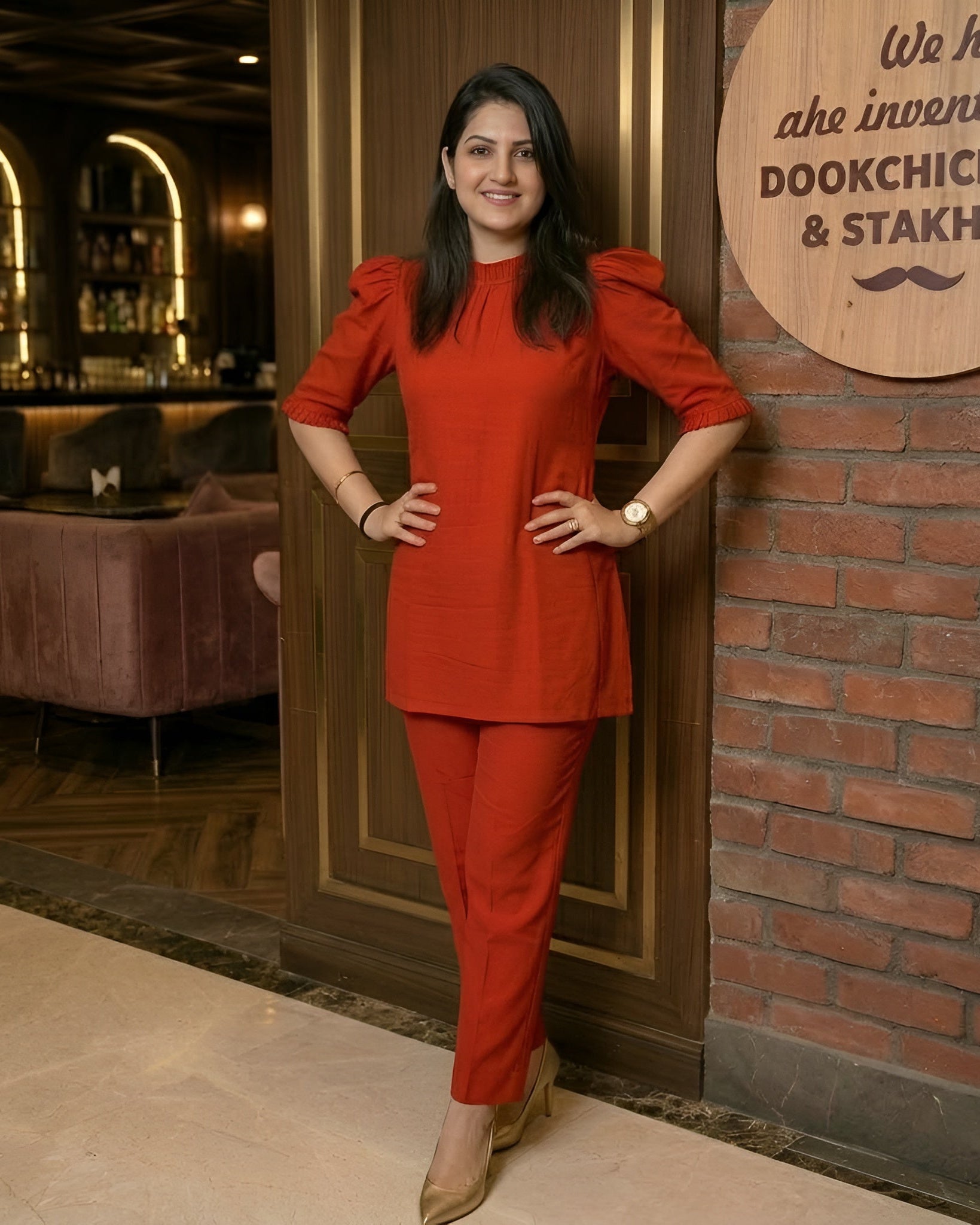 Woman in a red outfit posing in a restaurant setting with a decorative wall sign.