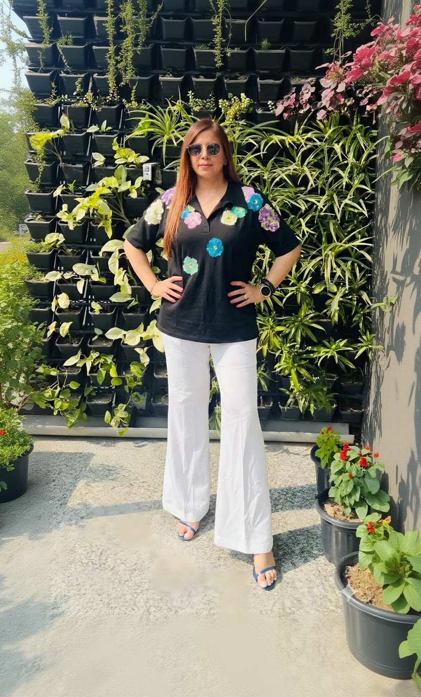 Woman posing in front of a plant wall wearing a black shirt with colorful designs and white pants.