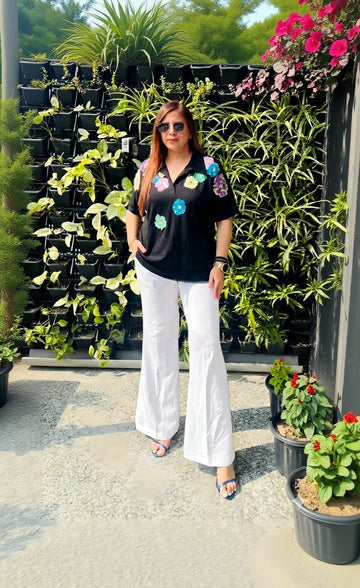 Woman wearing a black shirt with colorful floral patterns and white pants standing in front of potted plants.