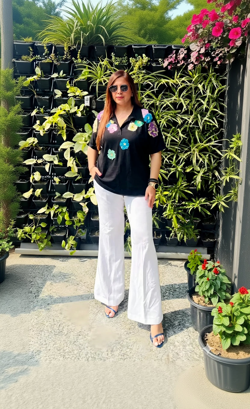 Woman wearing a black shirt with colorful floral patterns and white pants standing in front of potted plants.