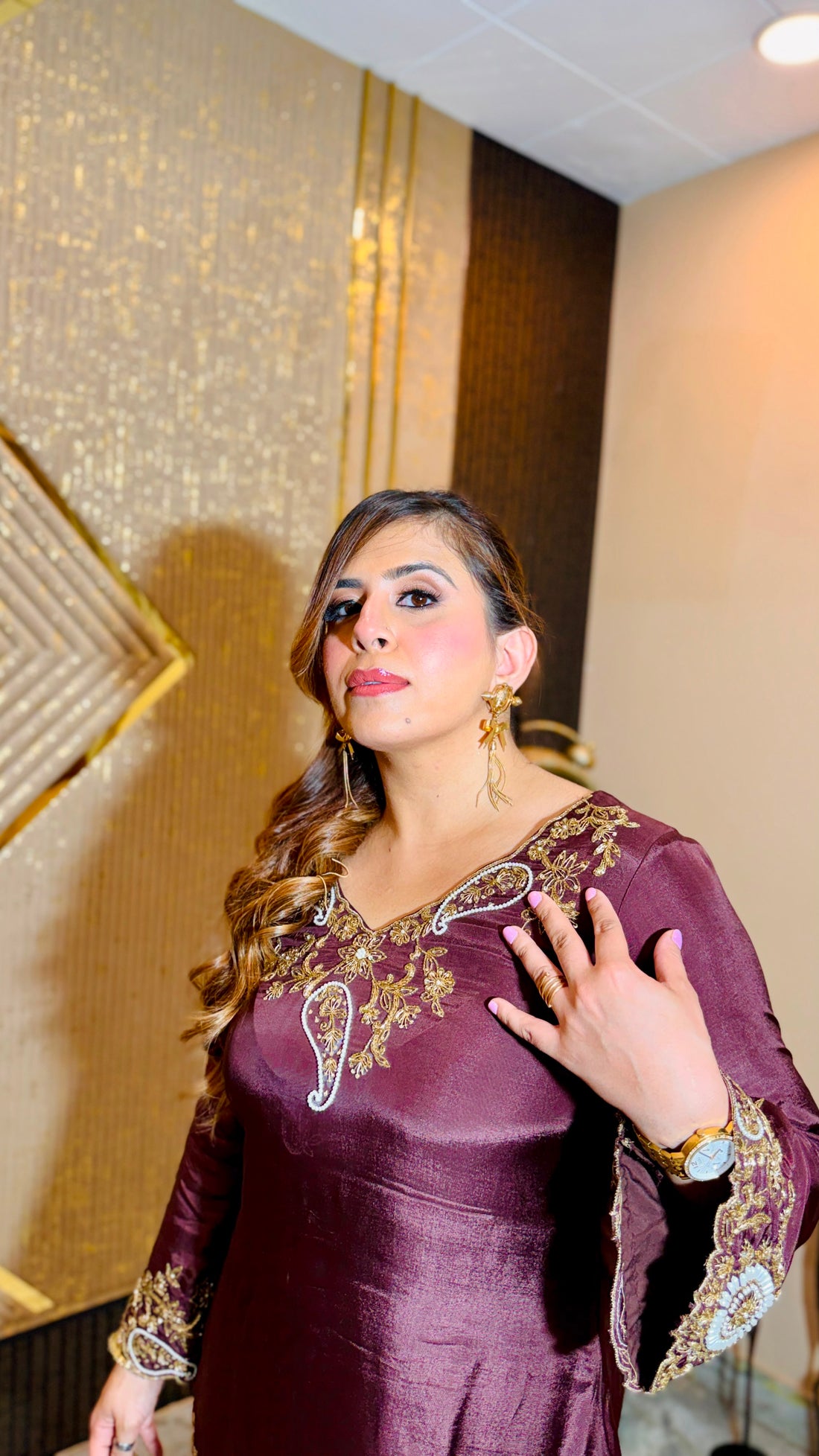 A woman models a deep maroon three-piece traditional outfit with gold embroidery, standing in an indoor setting with a patterned beige wall and decorative wooden furniture.