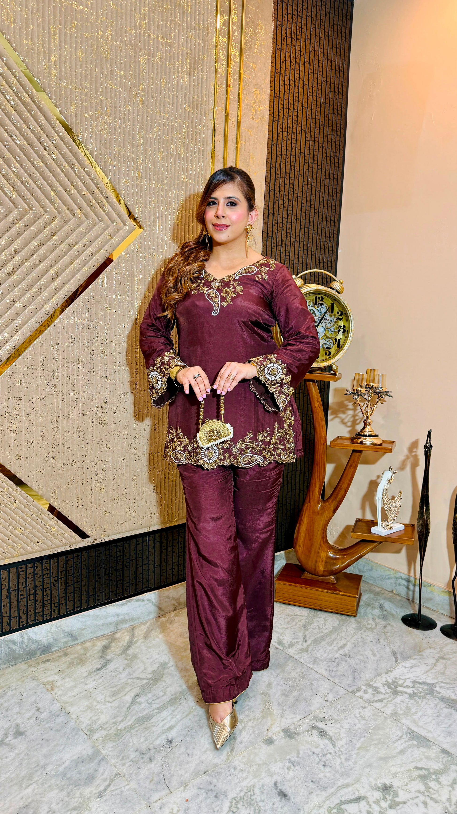 A woman models a deep maroon three-piece traditional outfit with gold embroidery, standing in an indoor setting with a patterned beige wall and decorative wooden furniture.
