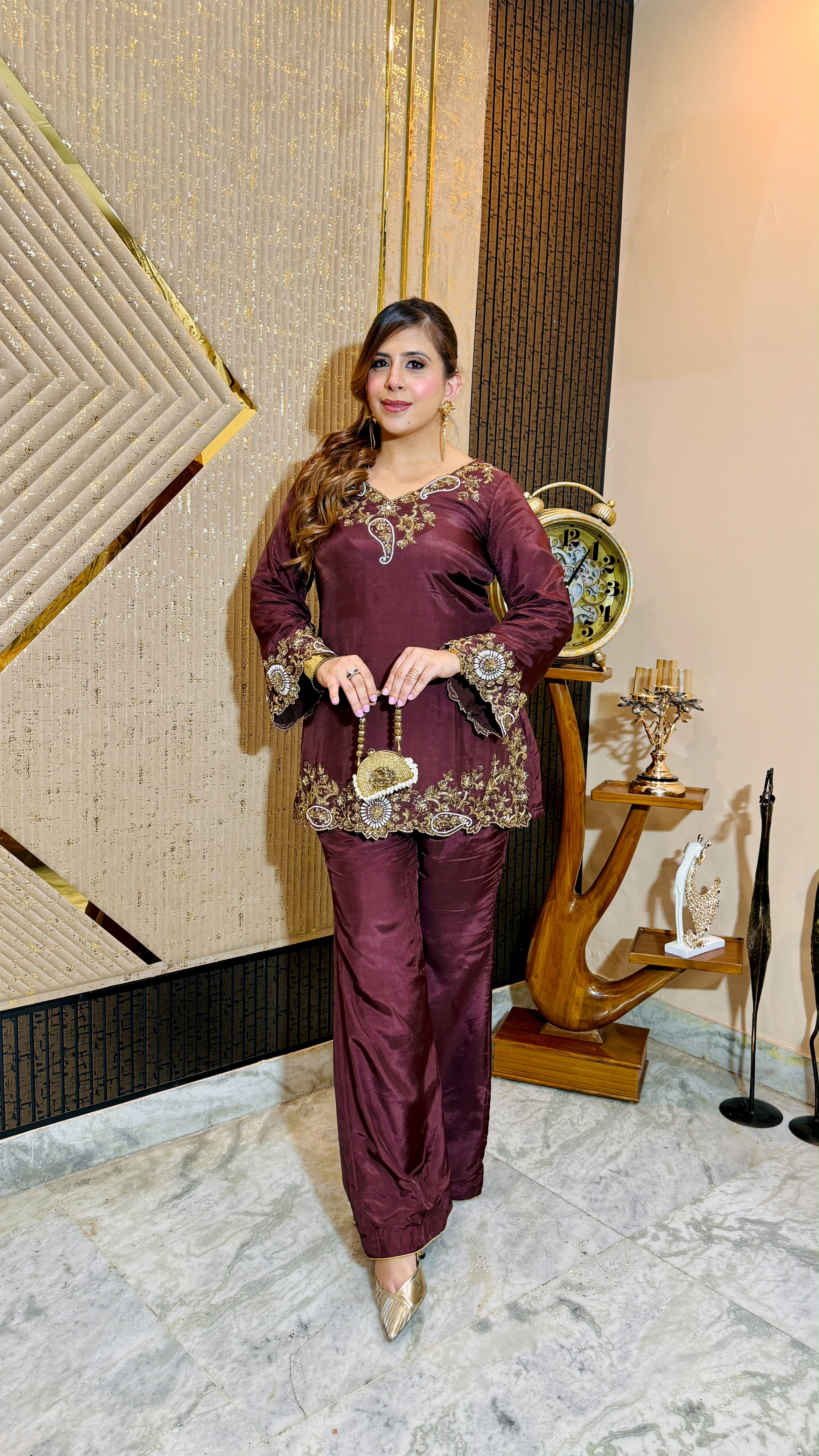 A woman models a deep maroon three-piece traditional outfit with gold embroidery, standing in an indoor setting with a patterned beige wall and decorative wooden furniture.
