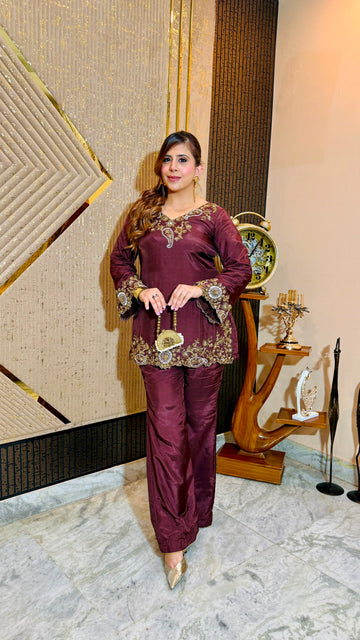 A woman models a deep maroon three-piece traditional outfit with gold embroidery, standing in an indoor setting with a patterned beige wall and decorative wooden furniture.