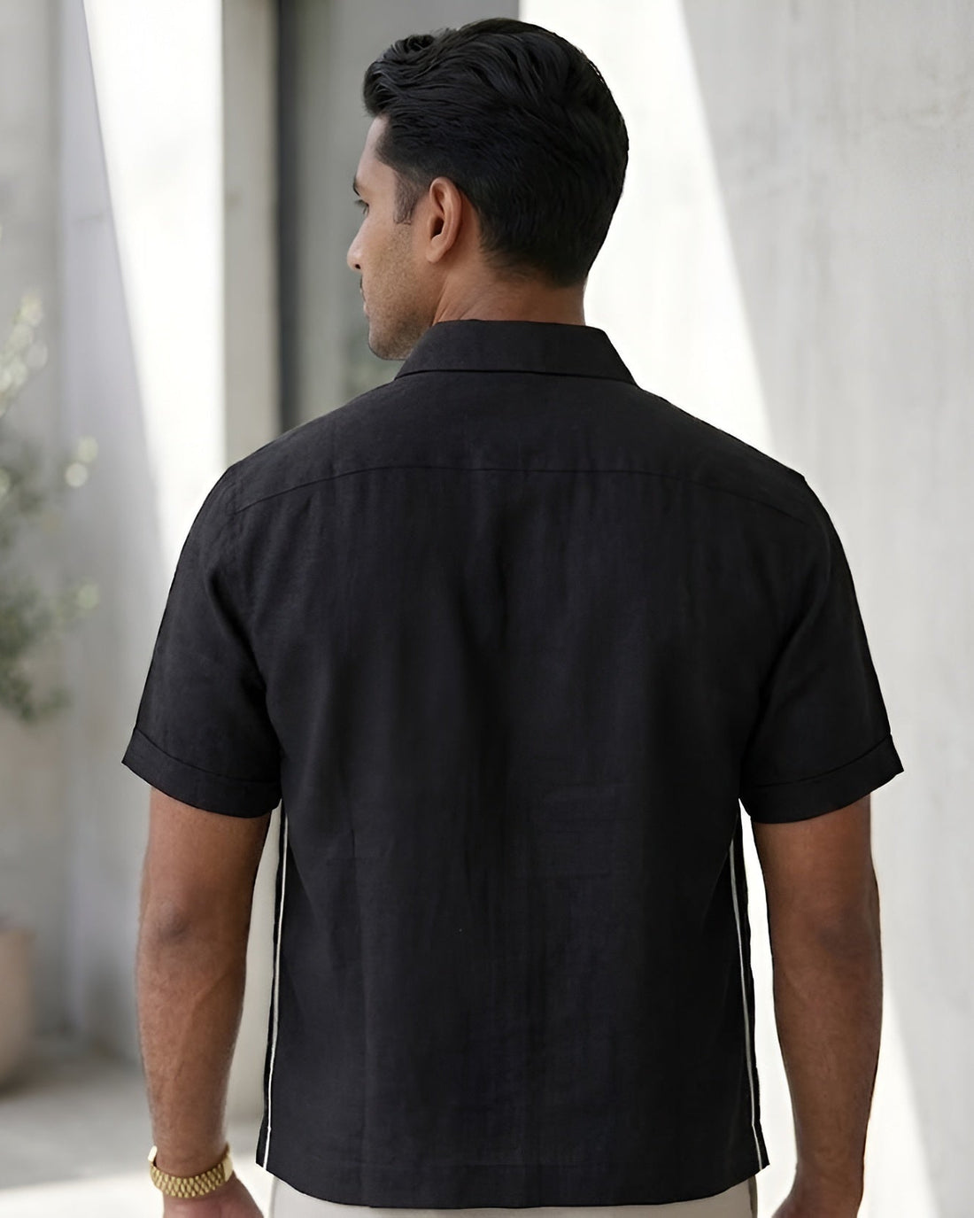 Man wearing a black embroidered shirt and white pants standing in a modern indoor setting with plants.