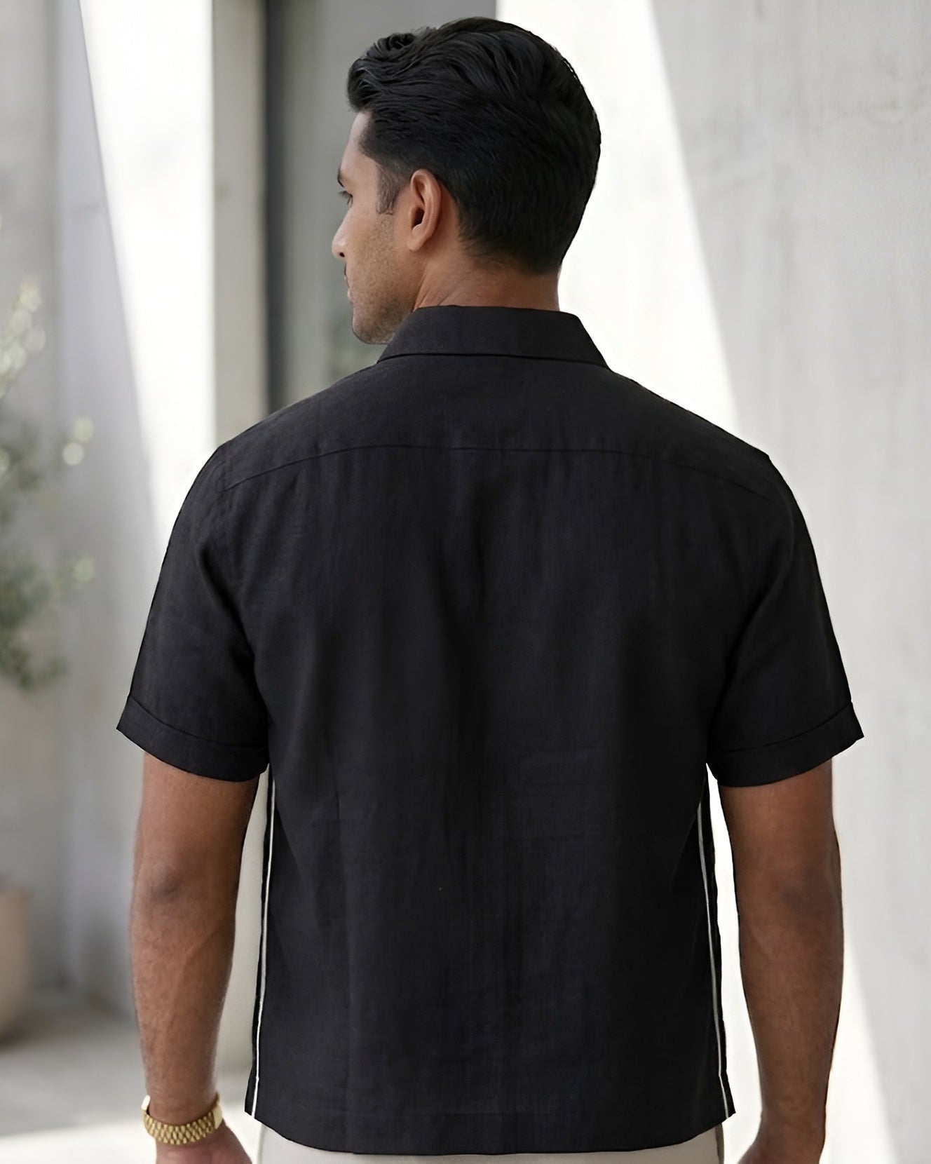 Man wearing a black short-sleeve shirt standing in a room with light gray walls and a plant.