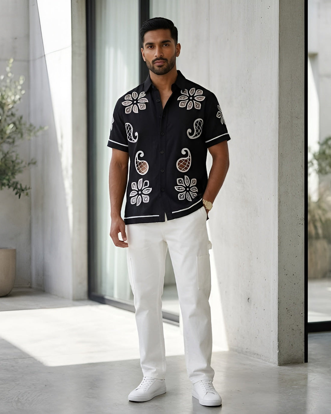 Man wearing a black embroidered shirt and white pants standing in a modern indoor setting with plants.