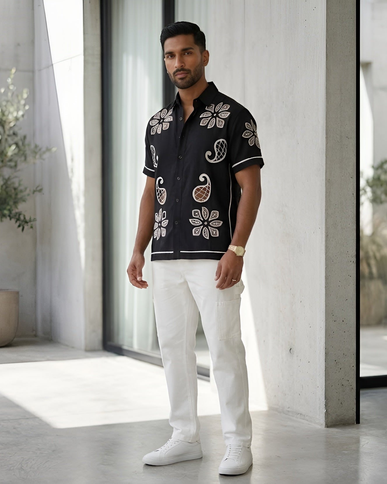 Man wearing a black floral shirt and white pants standing in a modern indoor setting with plants.