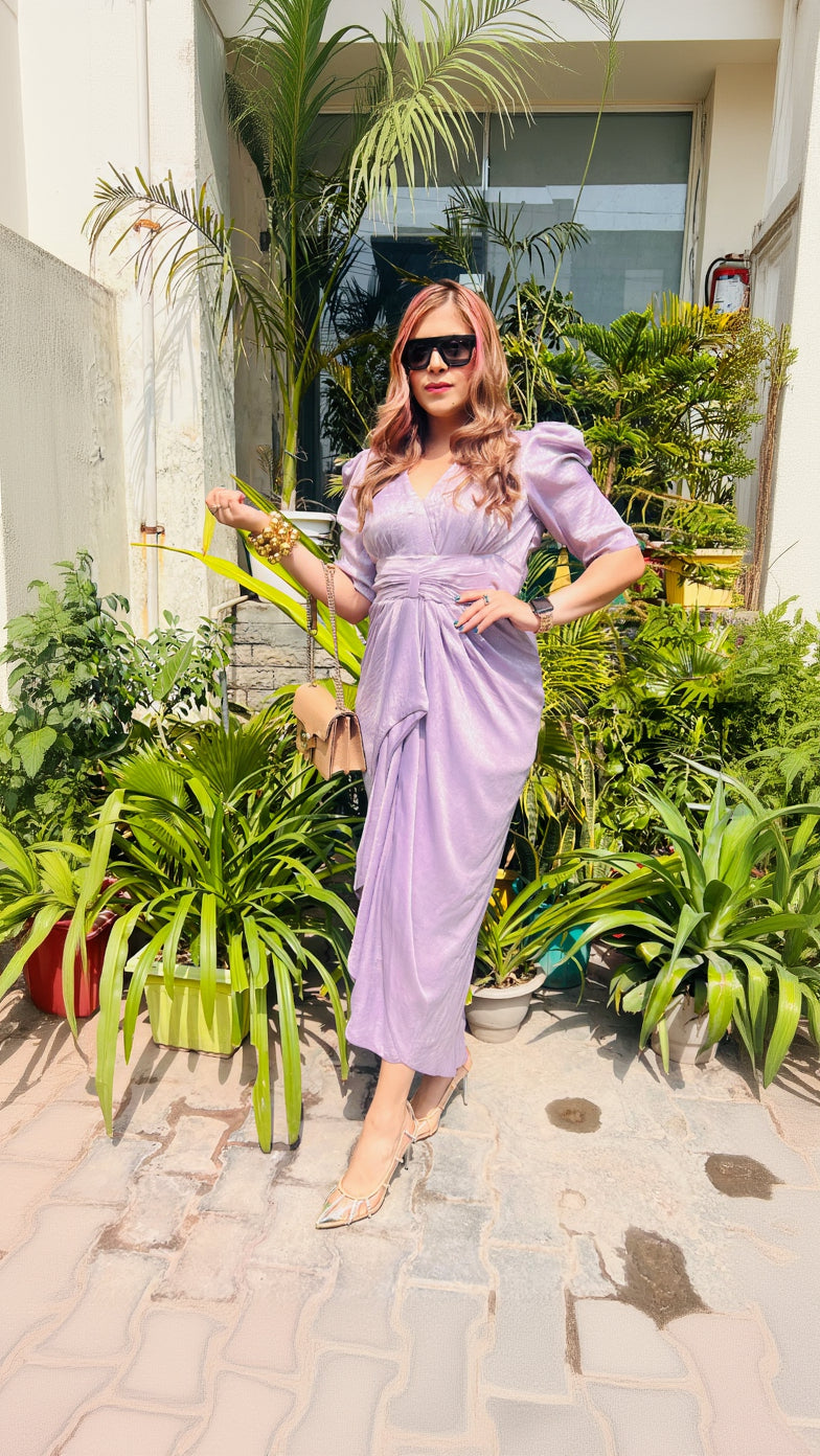 A person wearing a lilac-colored, metallic foil moss fabric drape with a plunging V-neck and elbow-length puffy sleeves, standing outdoors among plants.