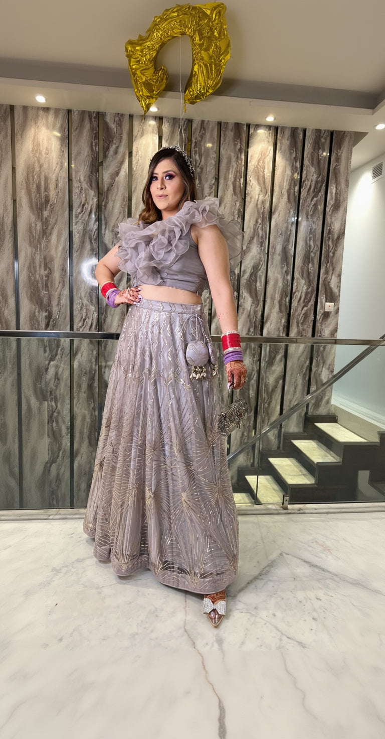 A woman wearing a grey net lehenga with embroidered details and a flared design, paired with a ruffle styled blouse, standing near a staircase.