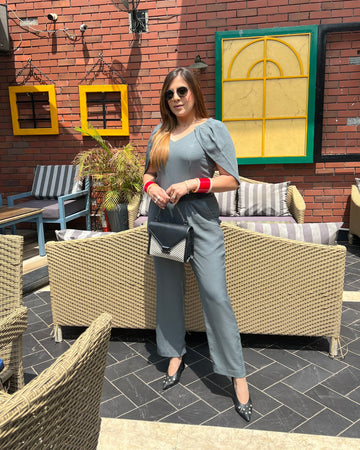 A person wearing a grey jumpsuit with sleeve detailing, standing in a outdoor seating area.
