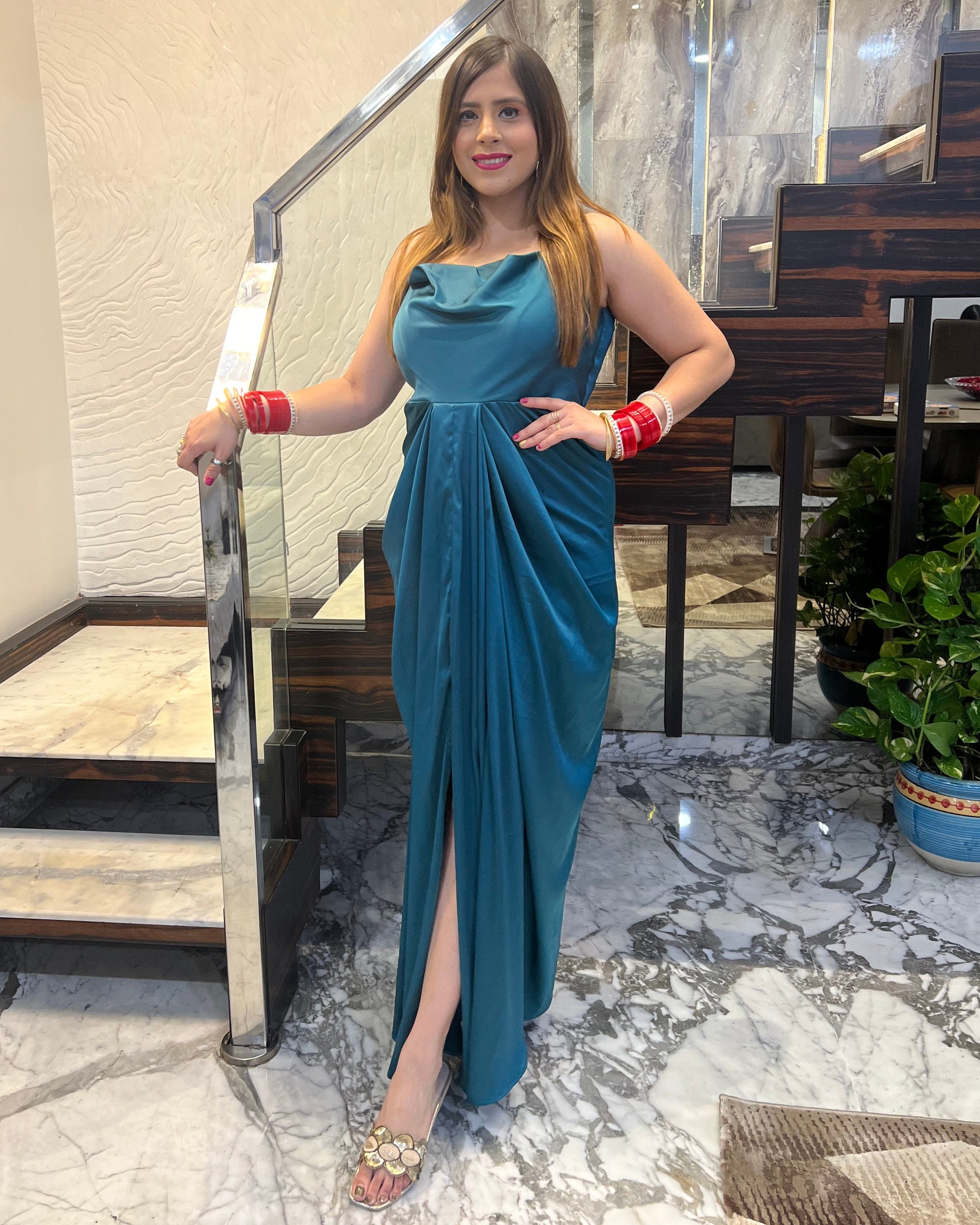 A woman wearing a teal green drape maxi dress with a cowl neckline and noodle straps, standing indoors.