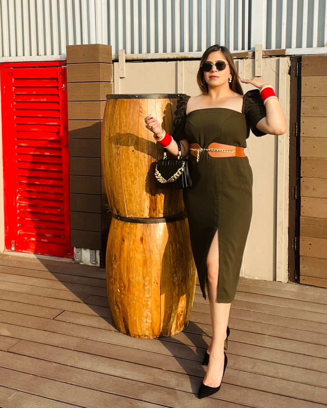 A woman standing on a wooden deck wearing an olive green solid midi dress with a belt, sunglasses, and high heels. She has accessorized with multiple bracelets and a watch.