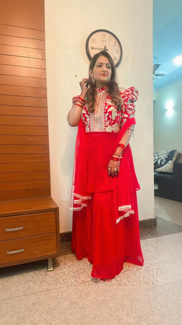 A woman wearing a traditional red Sharara suit with embroidered work, consisting of a peplum top and a long skirt, posing for the camera.