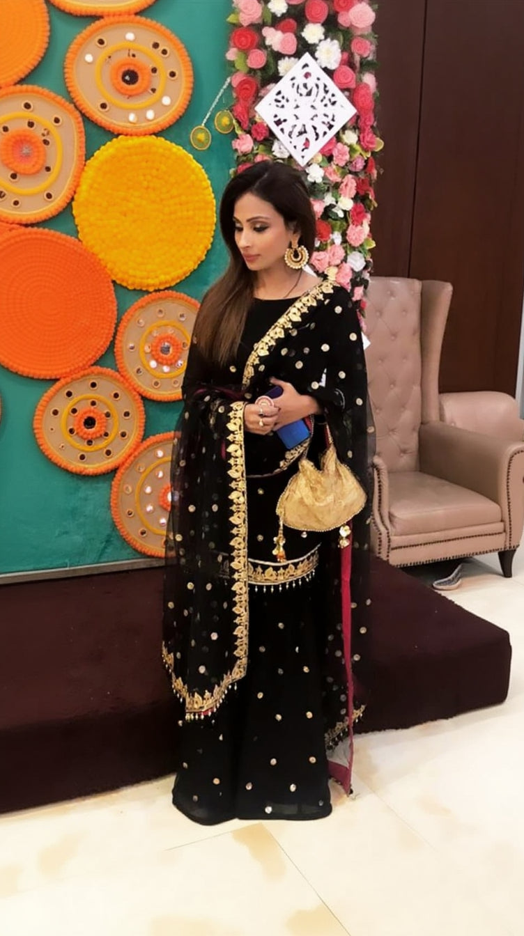 A woman wearing a black embroidered traditional suit with a dupatta, featuring floral and geometric patterns, attending a festive event.