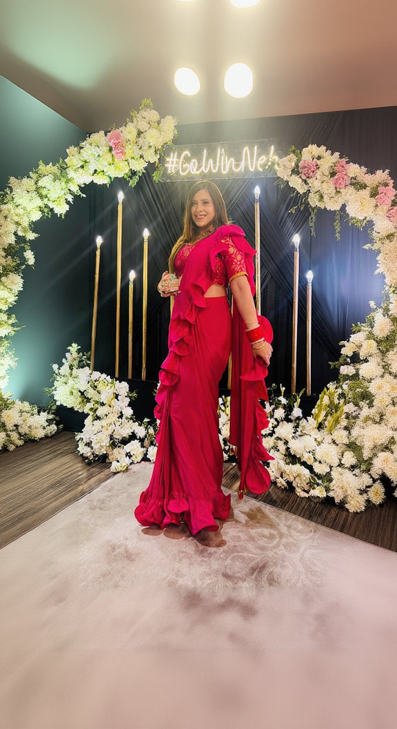 A woman posing in a hot pink ruffle saree with yellow detailing, featuring a front neck style, back neck style, and 3/4th regular sleeves with lace. The saree is made of pure Banarasi silk with French Crepe fabric.