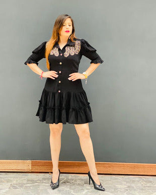 A woman standing against a grey background, wearing a black feather shirt dress with short puffed sleeves and knee-length, paired with heels.